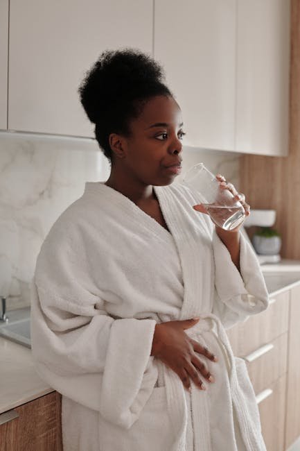 Woman stretches and drinks water by a sunlit kitchen window.